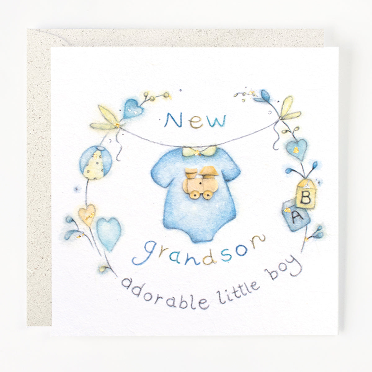A greeting card with a hand drawn illustration of baby clothing and accessories, with the text 'New grandson adorable little boy' on it, and a envelope behind it.