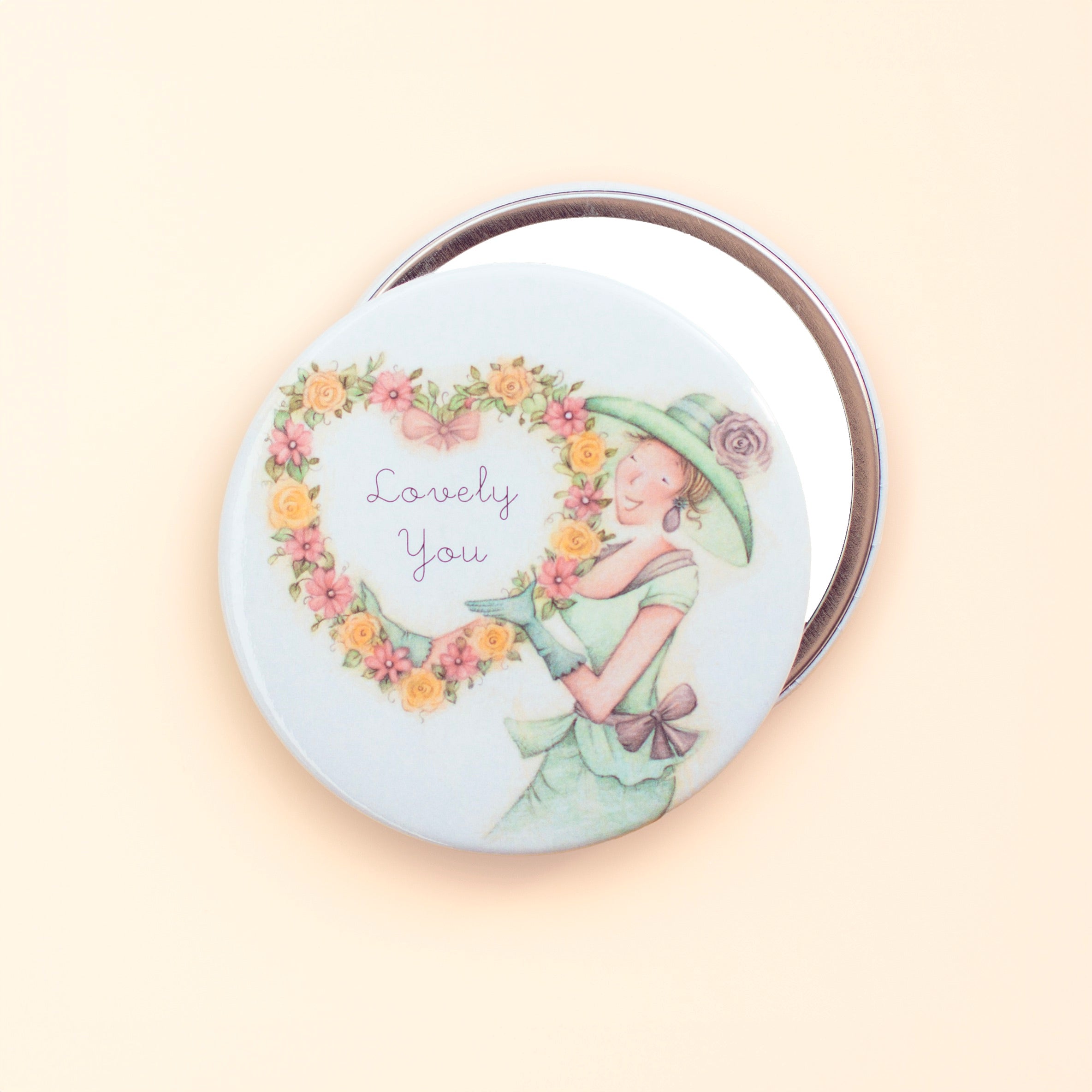 Round pocket mirror with an illustration of a woman in a green dress and a hat, surrounded by a floral border, with the text 'Lovely You' at the top.
