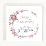 A greeting card with a floral wreath design and the text 'Happy Anniversary' in the center. There are two birds perched on hearts.